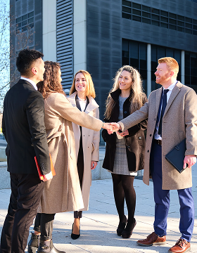 A group of five professionals stand outdoors in business attire, shaking hands and smiling. The setting conveys friendliness and a successful business meeting.